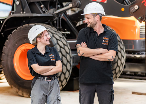 Zwei Bauarbeiter mit weißen Schutzhelmen und schwarzen Hemden stehen vor einer großen Maschine und lächeln sich mit verschränkten Armen an. Sie scheinen ein freundliches Gespräch in einer industriellen Umgebung zu führen.