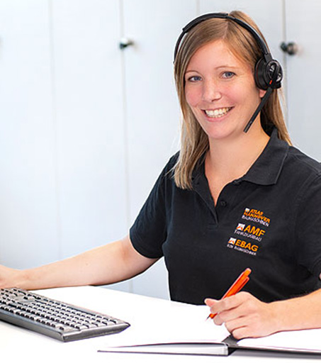 Kontakt Eine Frau mit Headset und schwarzem Poloshirt sitzt lächelnd an einem Schreibtisch, tippt auf einer Tastatur und hält einen Stift über ein aufgeschlagenes Notizbuch.