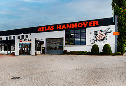 Ein großes Gebäude mit ATLAS HANNOVER in fetten orangefarbenen Buchstaben auf der Fassade, einer Wandmalerei mit einer Hand, die Werkzeuge hält, Glastüren und einer gepflasterten Fläche davor. Auf der rechten Seite ist etwas Grün zu sehen.