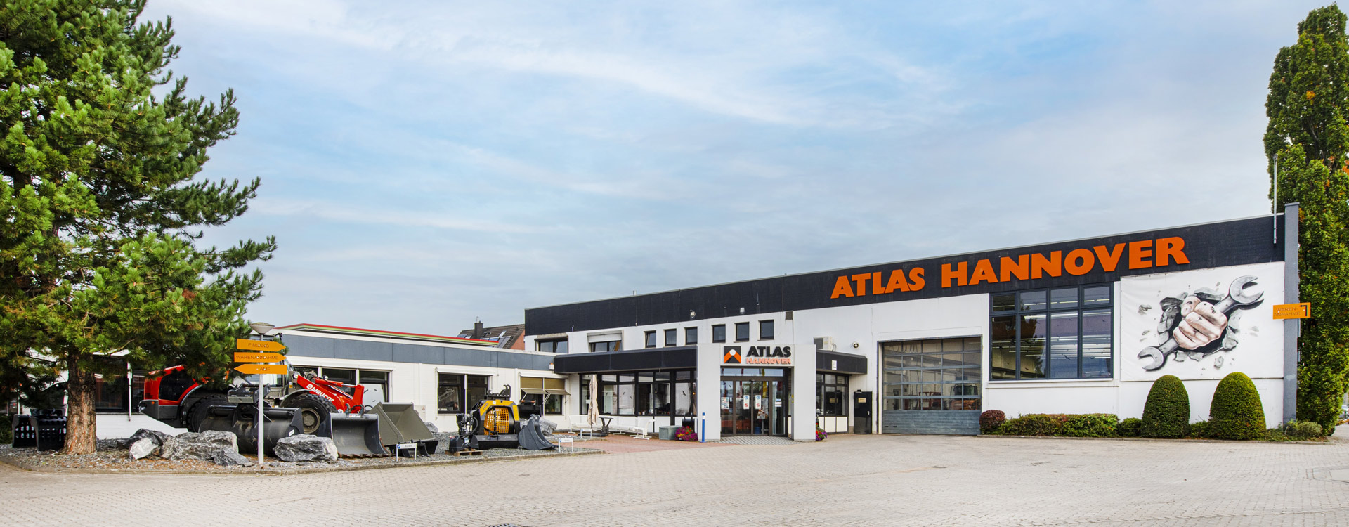 Das Bild zeigt das Äußere des ATLAS HANNOVER Gebäudes mit großen orangefarbenen Schildern. Draußen sind Baumaschinen ausgestellt, und auf der rechten Seite des Gebäudes befindet sich eine Wandmalerei mit einer mechanischen Klaue. Auf der linken Seite sind Bäume zu sehen.