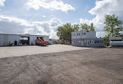 Ein großer offener Kiesplatz mit einem zweistöckigen Bürogebäude auf der rechten Seite, einer Metallgarage oder Werkstatt auf der linken Seite, einem roten Lastwagen, der in der Nähe der Werkstatt geparkt ist, und Bäumen im Hintergrund unter einem teilweise bewölkten Himmel.
