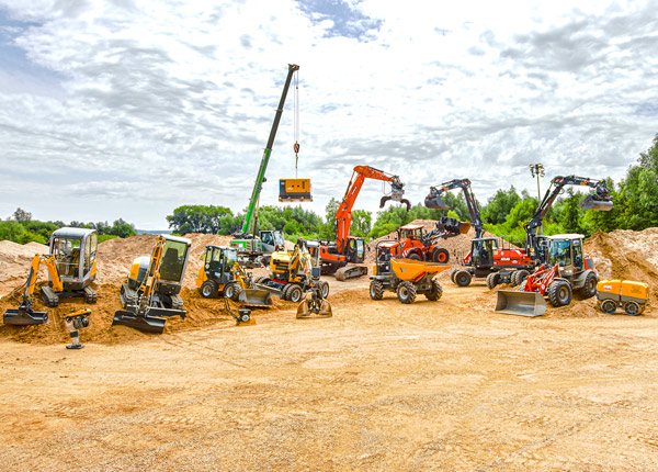 Eine Vielzahl von Baufahrzeugen und -geräten, darunter Bagger, Lader, Kräne und Muldenkipper, stehen auf einer unbefestigten Baustelle unter blauem Himmel mit vereinzelten Wolken und Sonnenlicht.