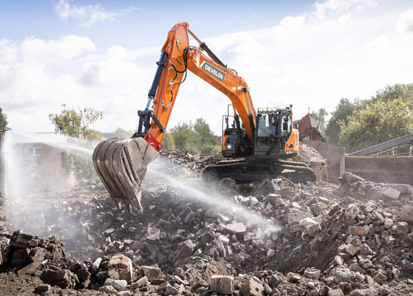 Ein orangefarbener Bagger bewegt an einem sonnigen Tag Schutt auf einer Abbruchbaustelle. Im Hintergrund sind Bäume und teilweise abgerissene Gebäude zu sehen.