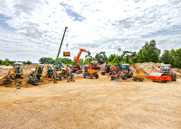 Eine Vielzahl von Baufahrzeugen und -geräten, darunter Bagger, Lader, Kräne und Muldenkipper, stehen auf einer unbefestigten Baustelle unter blauem Himmel mit vereinzelten Wolken und Sonnenlicht.