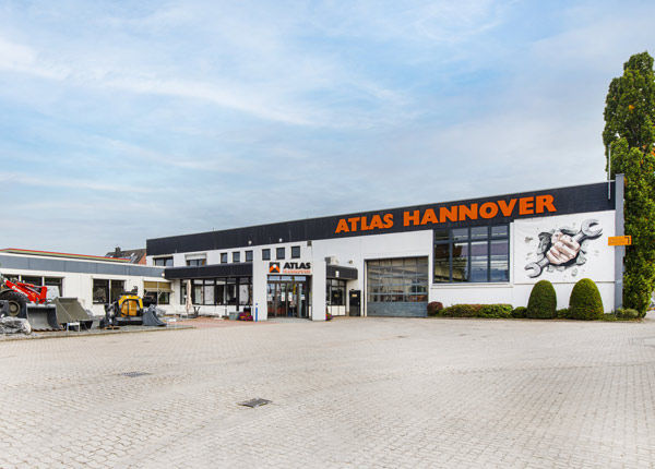 Ein Geschäftsgebäude mit der Aufschrift ATLAS HANNOVER mit einer großen Wandmalerei einer Hand, die Schraubenschlüssel hält, an der Außenwand, mit einer gepflasterten Fläche und etwas Grün im Außenbereich.