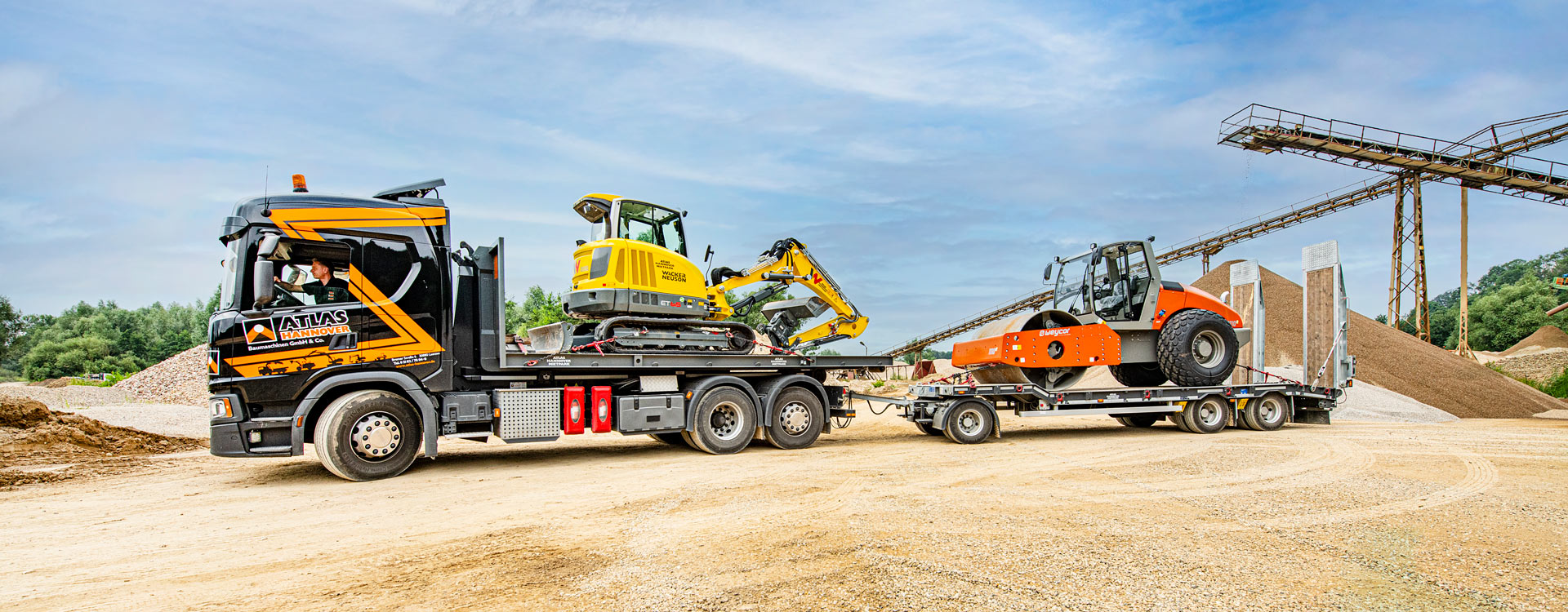 Ein Lkw mit Anhänger, der Baumaschinen, darunter einen gelben Bagger und eine orangefarbene Straßenwalze, transportiert, parkt auf einem unbefestigten Grundstück unter blauem Himmel.
