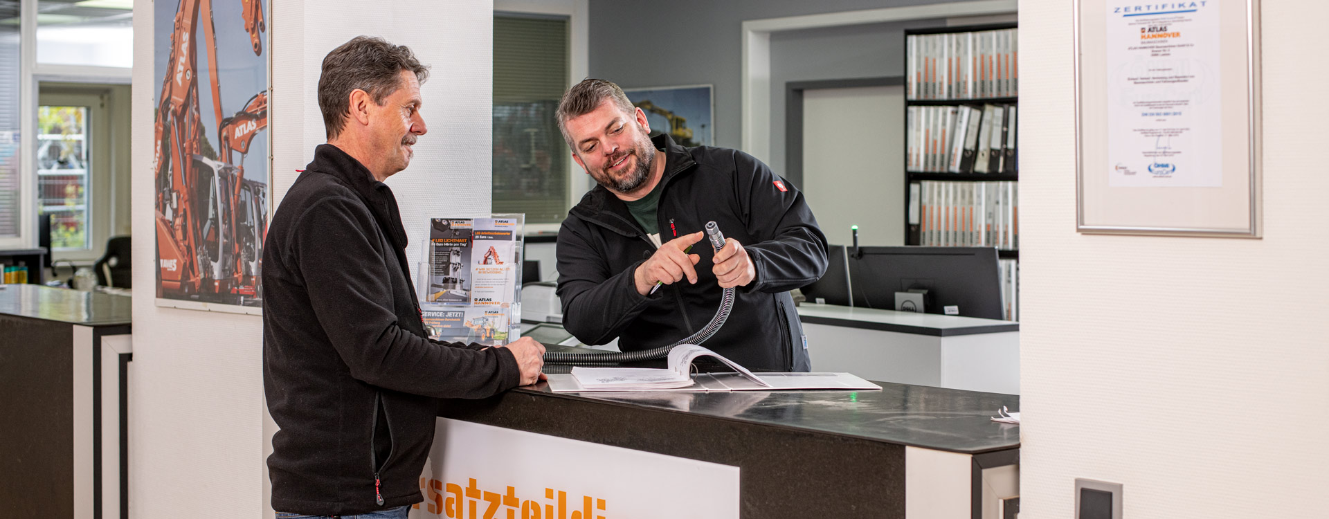 Zwei Männer stehen an einem Serviceschalter in einem Büro. Ein Mann zeigt lächelnd auf ein Kabel, der andere schaut auf einen aufgeschlagenen Katalog. Im Hintergrund sind Regale mit Aktenordnern und einer gerahmten Urkunde zu sehen.