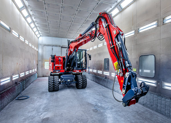 Ein roter Bagger mit einem langen Hydraulikarm steht in einer hell erleuchteten industriellen Lackierkabine mit grauen Wänden, Lüftungsschlitzen und Deckenbeleuchtung.