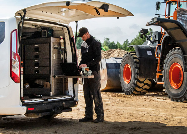 Ein Arbeiter in schwarzer Jacke und Mütze benutzt einen Laptop auf einem ausklappbaren Tisch im hinteren Teil eines Lieferwagens, der neben schweren Baumaschinen auf einer Baustelle geparkt ist.