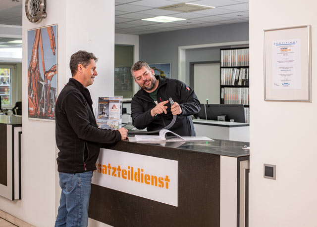 Zwei Männer stehen an einem Empfangstresen in einem Büro, schauen sich gemeinsam ein Dokument an und lächeln. Auf dem Schreibtisch steht ein Schild mit der Aufschrift "Ersatzteildienst". Im Hintergrund sind Bürogeräte und Akten zu sehen.
