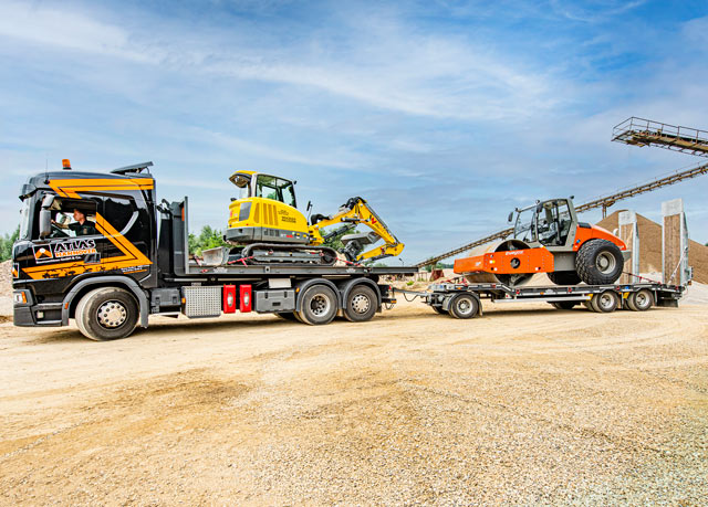 Ein Lkw mit Anhänger, der Baumaschinen, darunter einen gelben Bagger und eine orangefarbene Straßenwalze, transportiert, parkt auf einem unbefestigten Grundstück unter blauem Himmel.